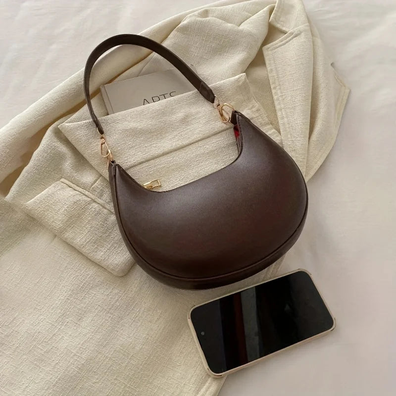 Luxury moon-shaped handbag in brown leather with a stylish handle, placed next to a smartphone