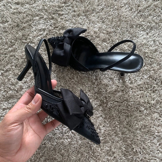Black mesh embroidered high heels with bow elegantly displayed by hand on a plush surface.