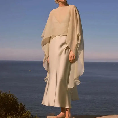Elegant women's chiffon overlay cape in beige, styled over a silky dress by VaultHaus against a coastal background